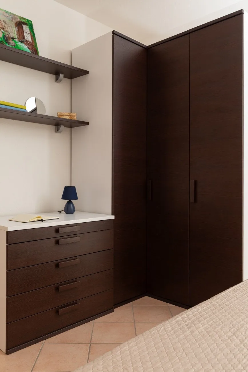 Una esquina de dormitorio con armario oscuro, cómoda y lámpara de mesa.