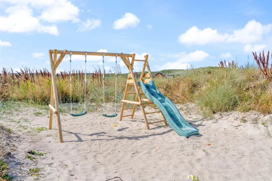 Un área de juegos al aire libre con columpios y un tobogán está sobre arena entre hierbas de dunas.