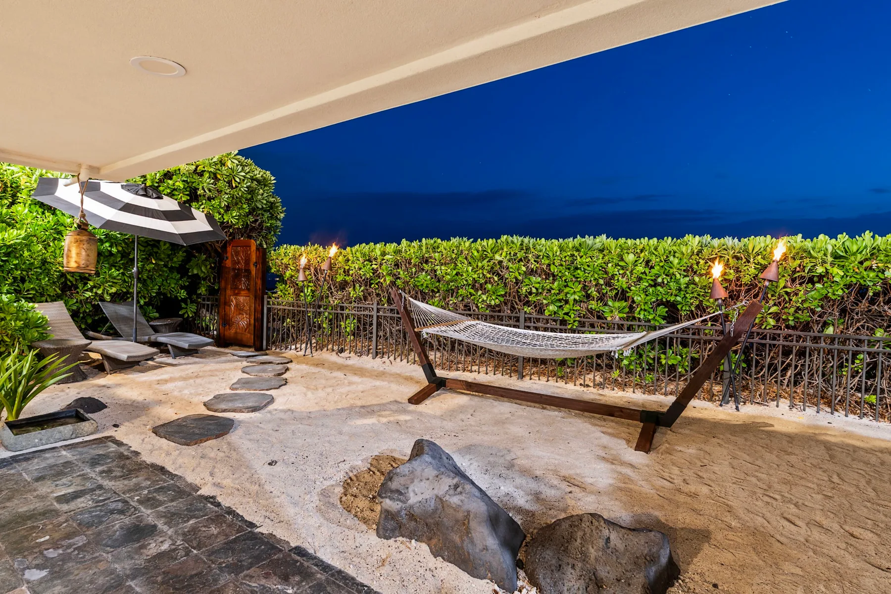 Un patio con una hamaca y sillones está junto a un seto verde y frondoso.