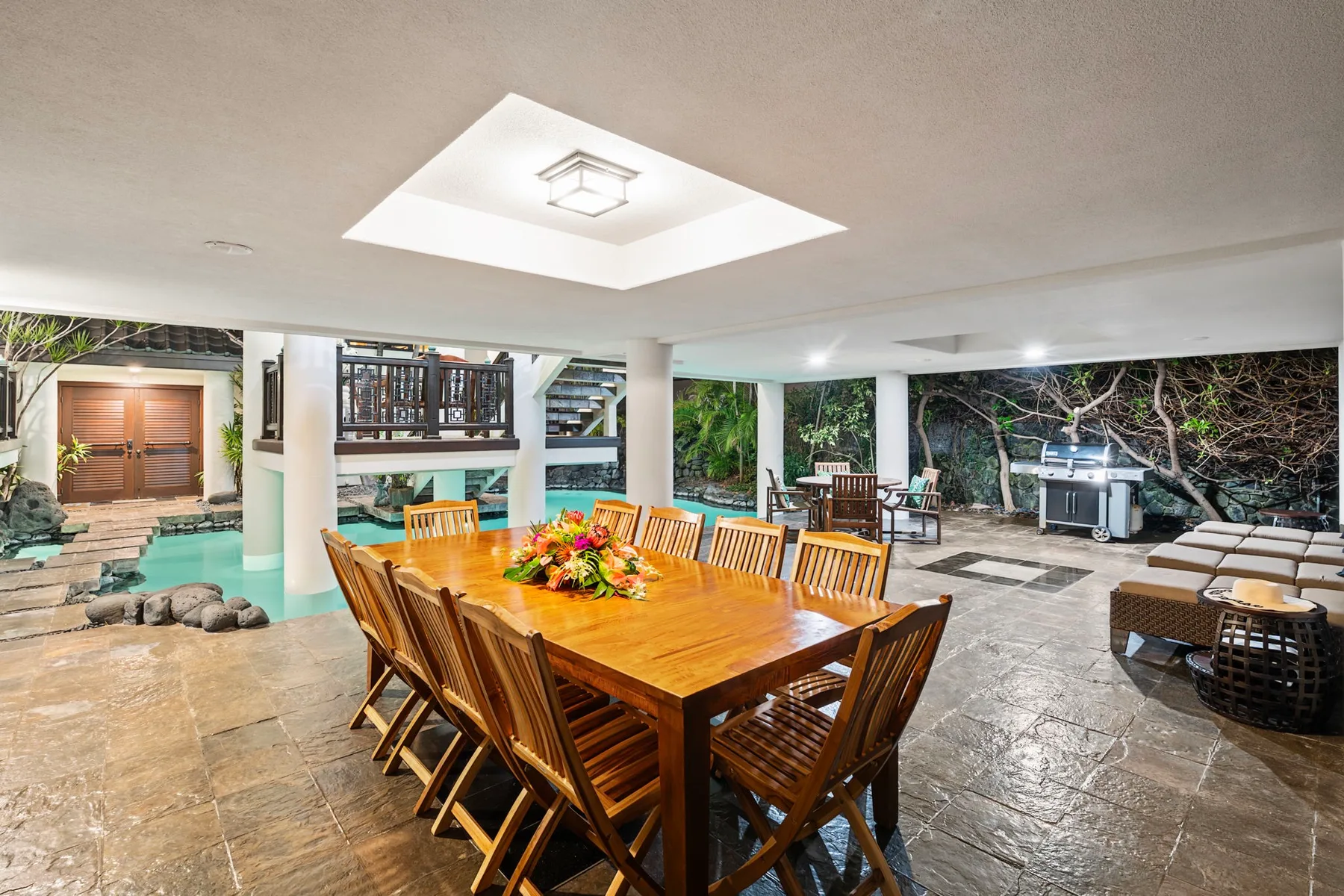 Una zona de comedor exterior cubierta tiene una gran mesa de madera rodeada de sillas.