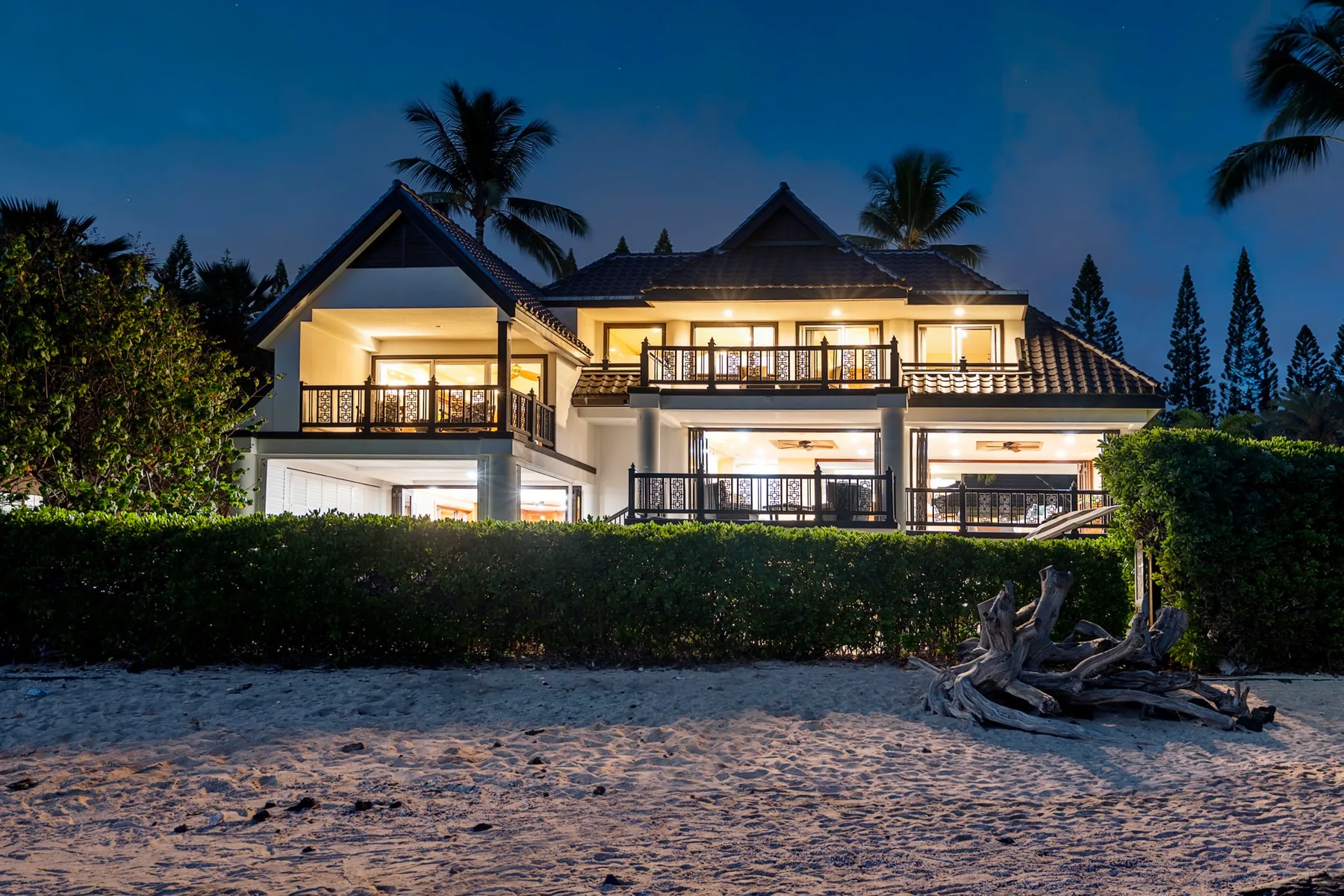 Una casa iluminada se alza detrás de un seto junto a una playa de arena al anochecer.