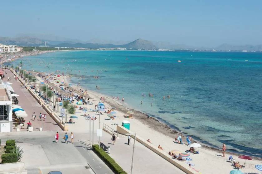 A crowded beach and seaside promenade stretch beside turquoise water.