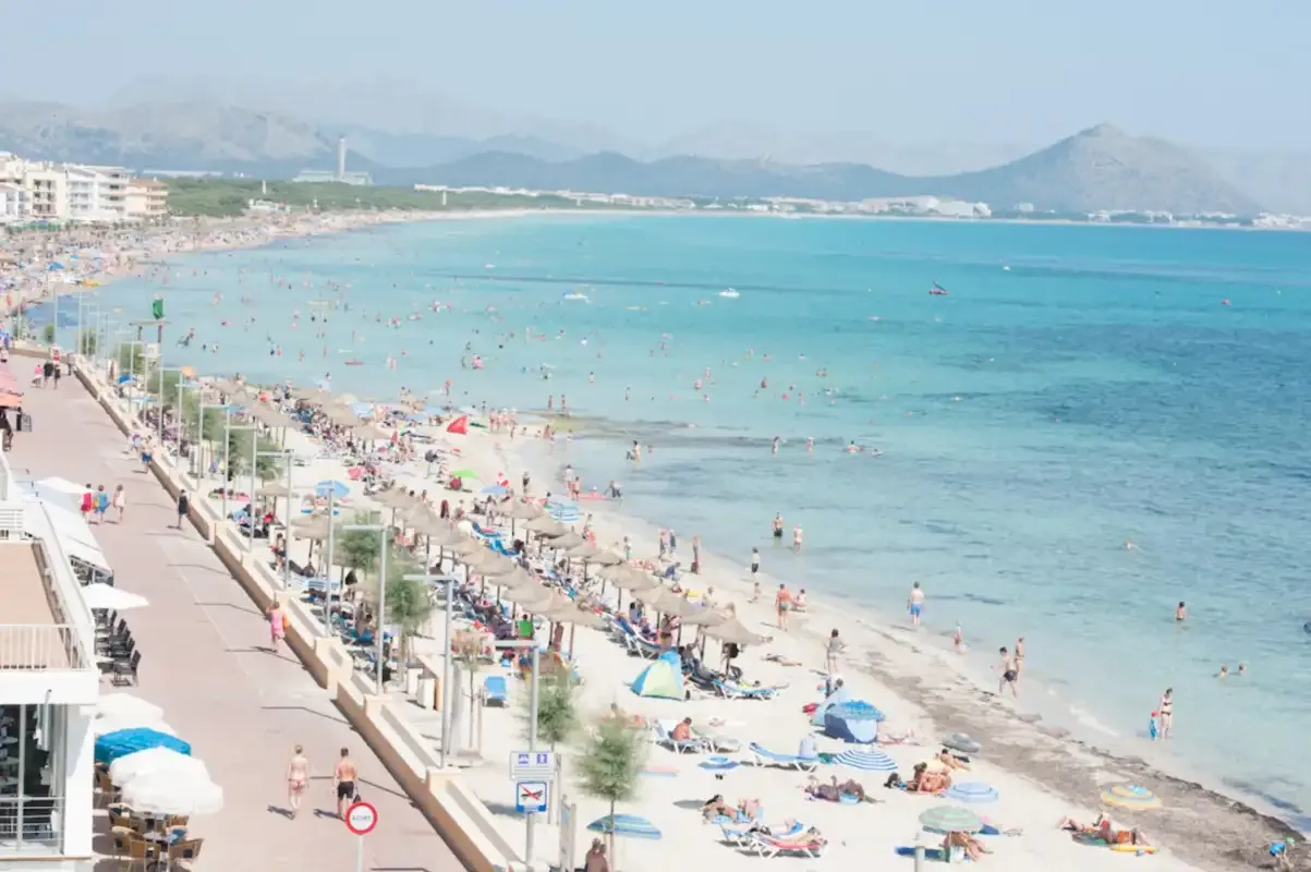 A crowded beach and seaside promenade beside clear blue water.