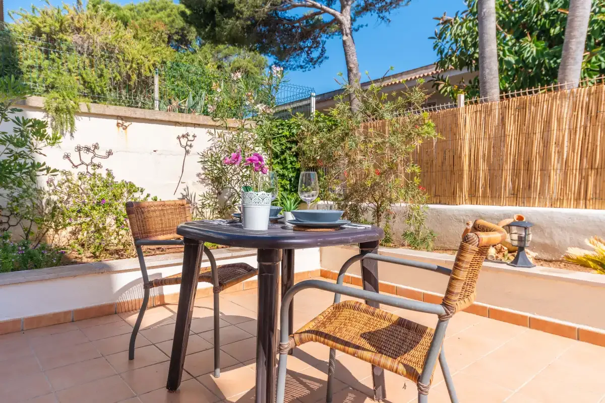 A sunny outdoor patio has a dining table, wicker seating, and potted flowers.