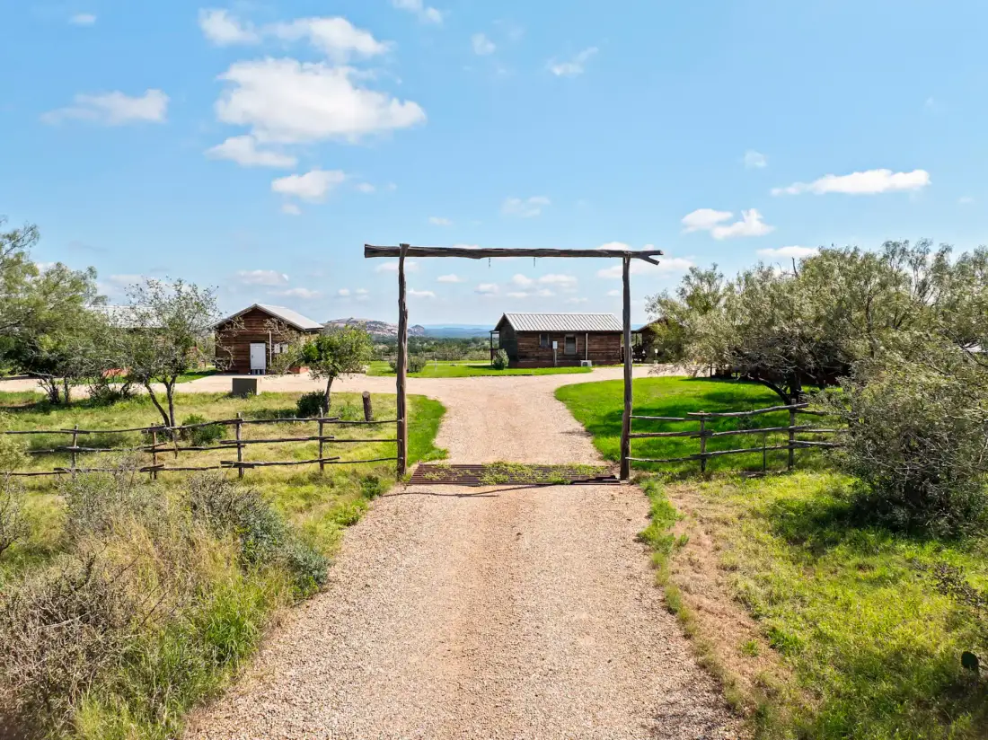 Gravel driveway leading to rustic cabins under a wooden arch, surrounded by greenery and under a blue sky with clouds.