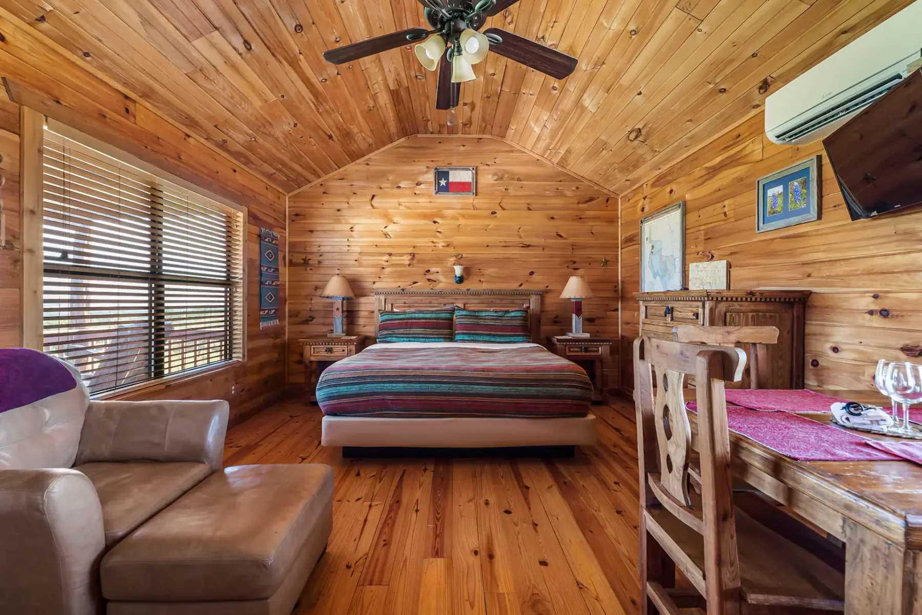 Cozy wooden cabin bedroom with a striped bedspread, armchair, dining table, ceiling fan, and air conditioning unit.
