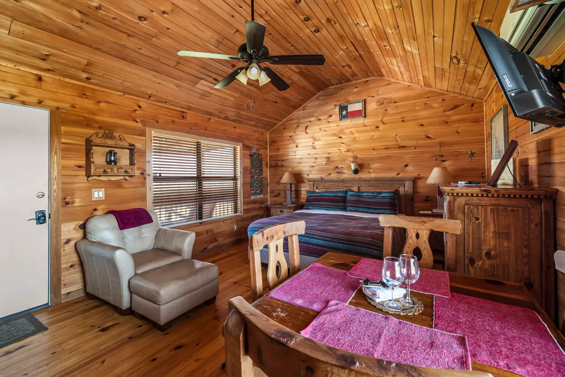 Cozy wooden cabin interior with a bed, armchair, dining table set with pink placemats, TV, ceiling fan, and window with blinds.