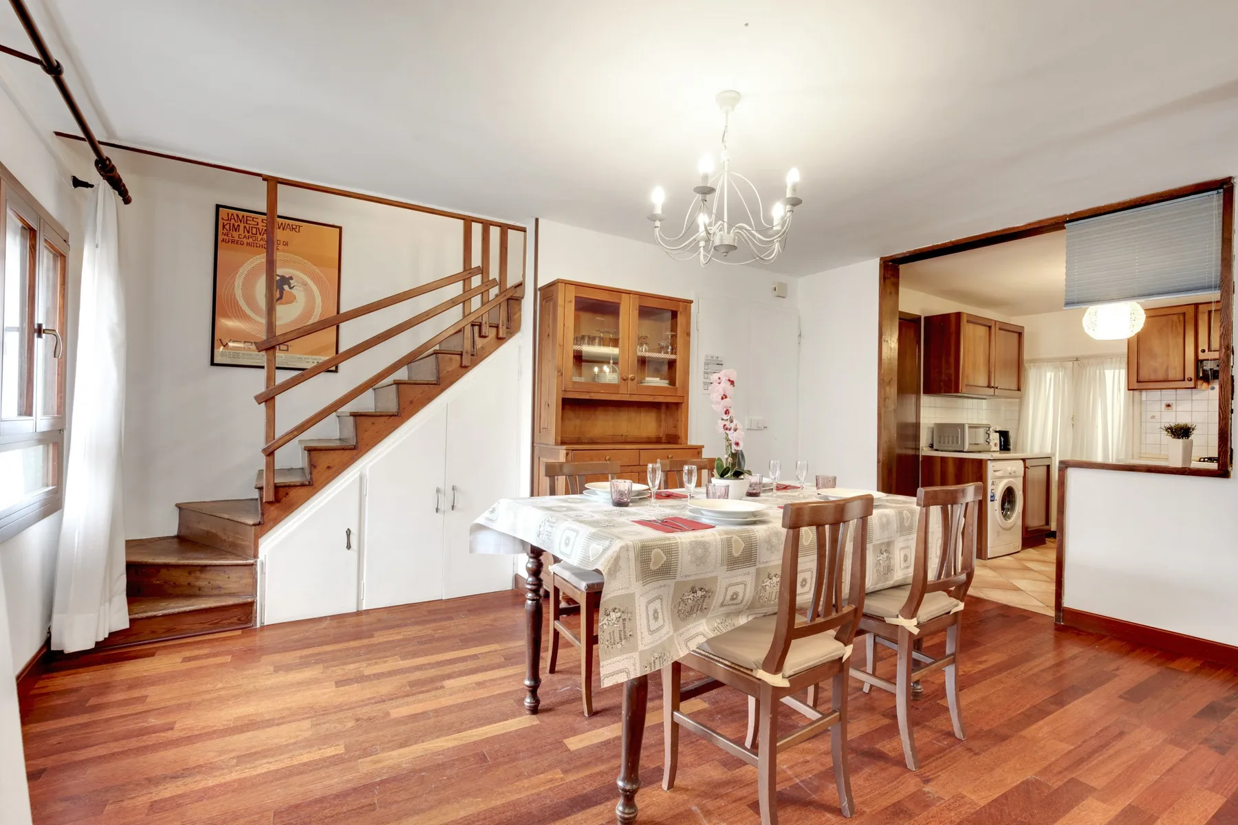 Comedor con la mesa puesta, junto a una escalera de madera y una cocina abierta.