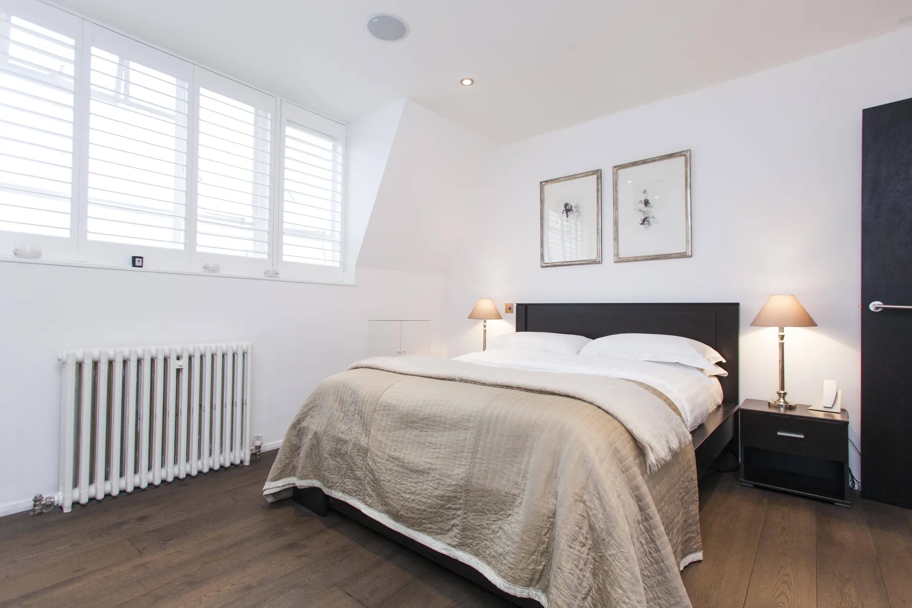 Un dormitorio luminoso con una cama bajo dos láminas enmarcadas y ventanas amplias.