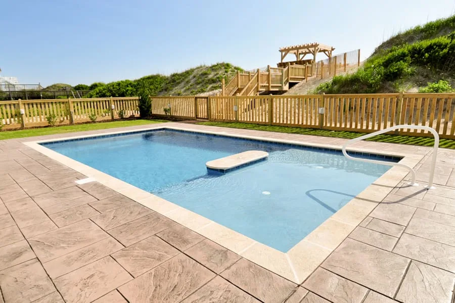 Una piscina al aire libre está rodeada por una terraza pavimentada y una cerca de madera.