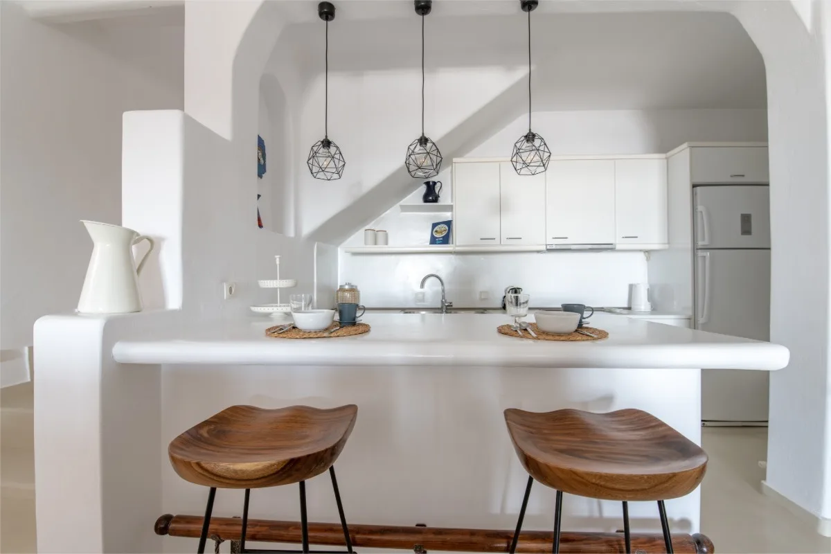 A bright kitchen with a white island, wooden seating, and pendant lights.
