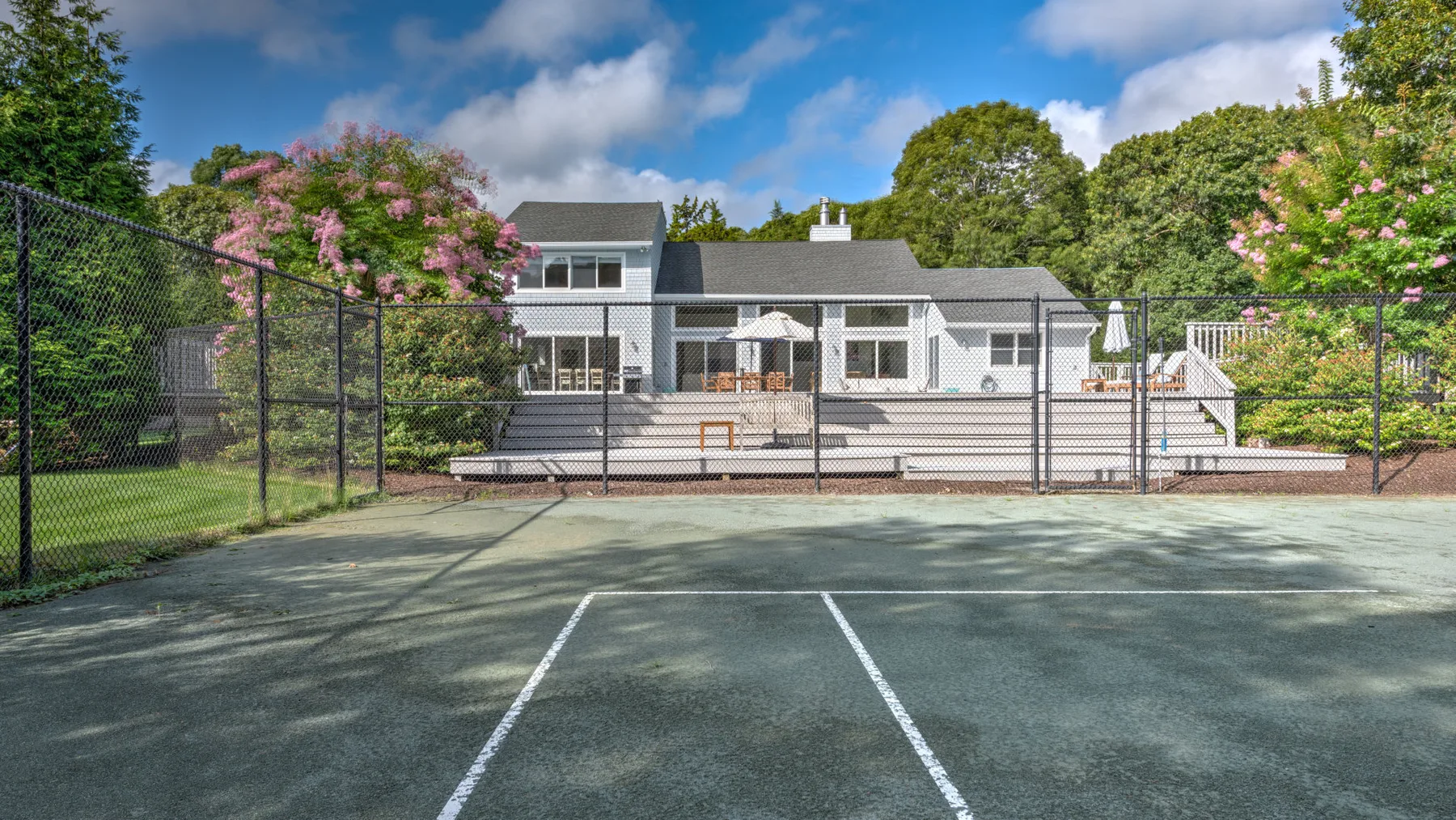 Una cancha de tenis al aire libre y cercada está frente a una casa y un patio.
