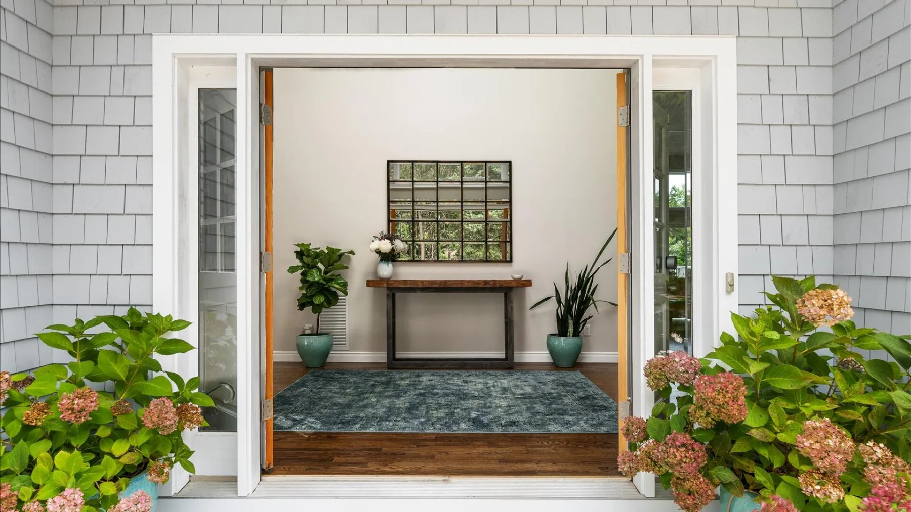 Una puerta abierta da a una habitación con una consola, plantas y una alfombra estampada.