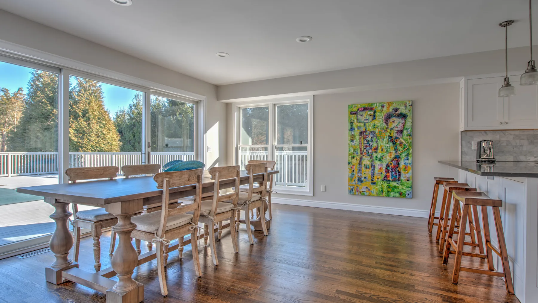 Un comedor luminoso con una mesa de madera junto a grandes ventanas y una pintura colorida en la pared.