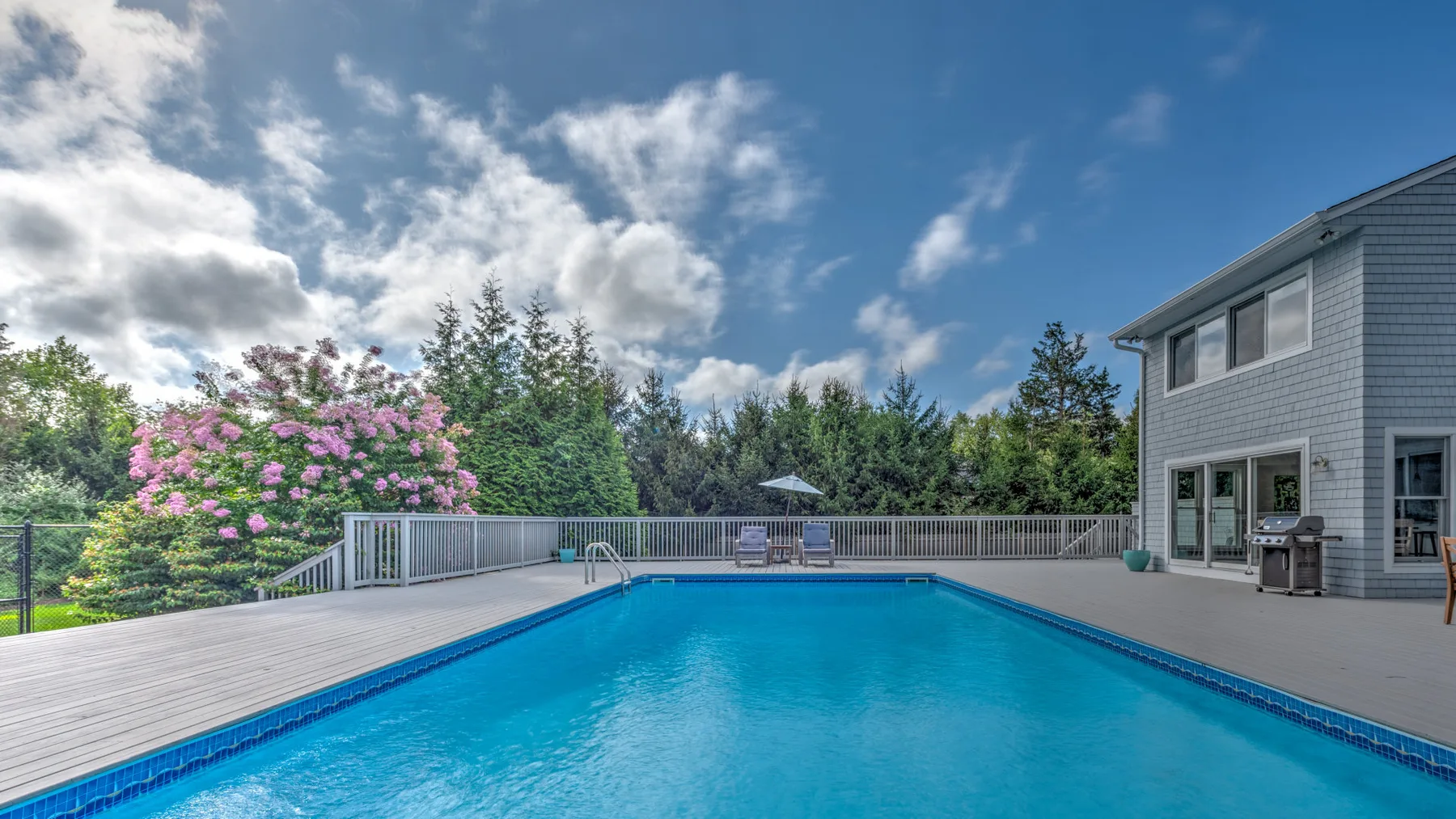 Una piscina al aire libre junto a una amplia terraza y una casa gris.