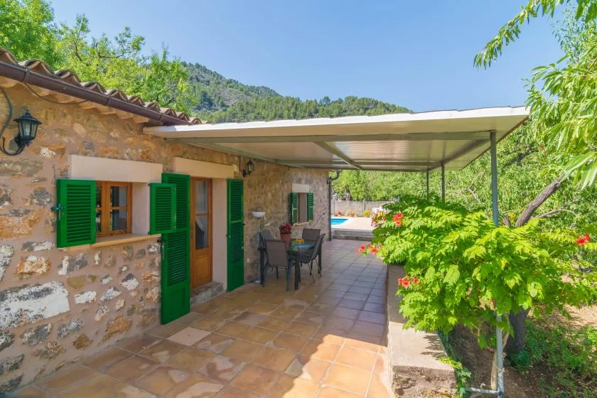 A shaded outdoor patio beside a stone building, with green shutters and garden plants.