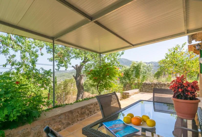 A covered outdoor patio with a glass table and views of greenery and hills.