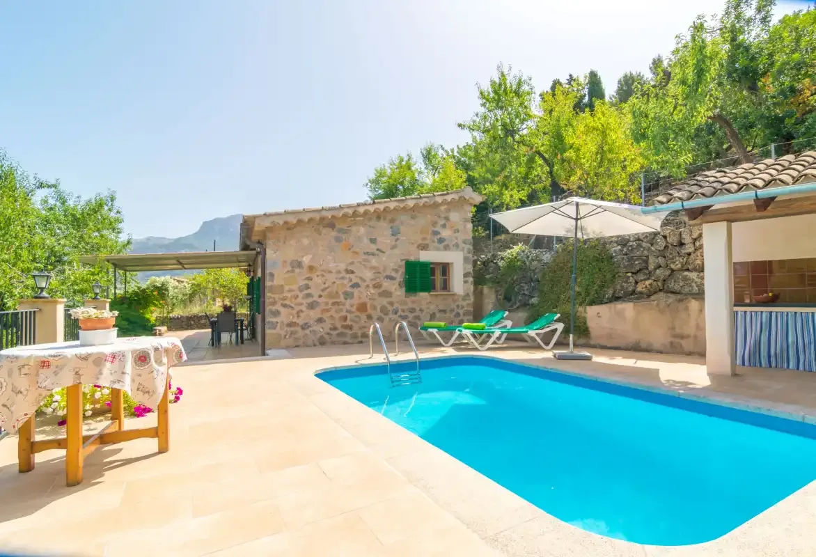 A swimming pool fills a sunny patio beside stone buildings and lounge seating.