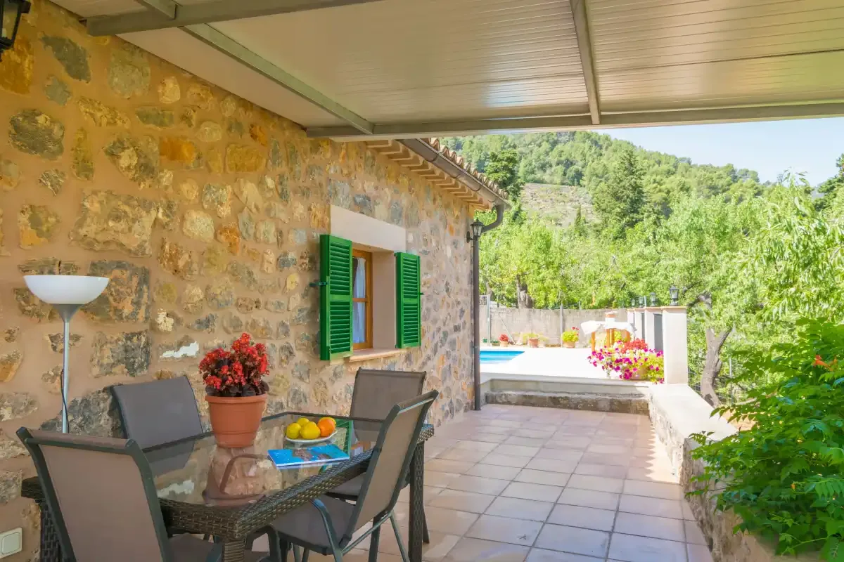 A covered stone patio with seating overlooks a garden and swimming pool.