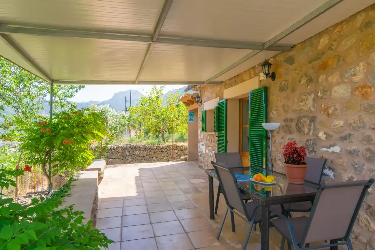 A covered outdoor patio with a dining table beside a stone wall and greenery.