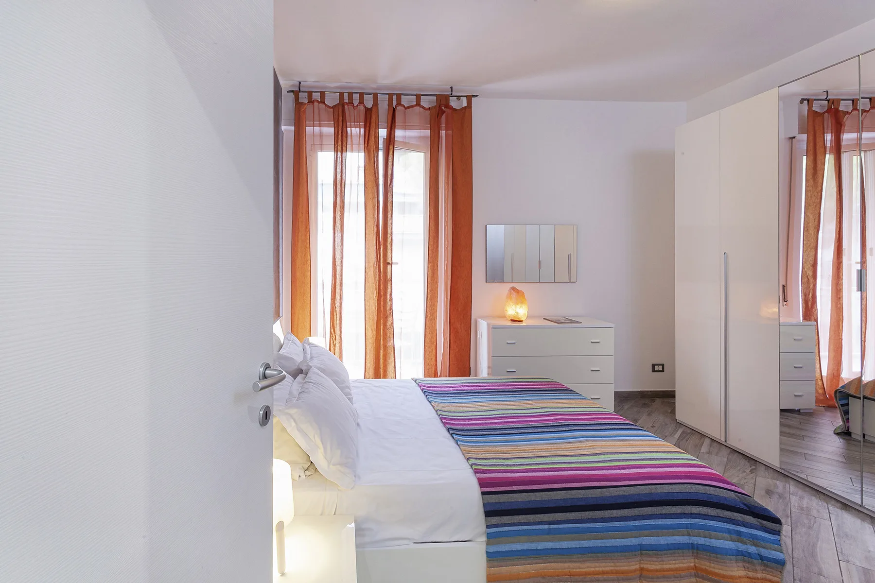A bedroom with a bed covered by a colorful striped blanket and orange curtains.