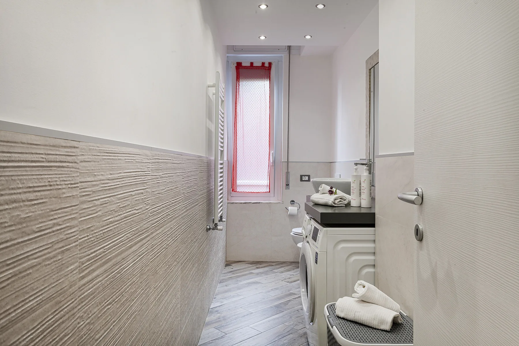 A narrow bathroom with a vanity and a tall window with a red curtain.