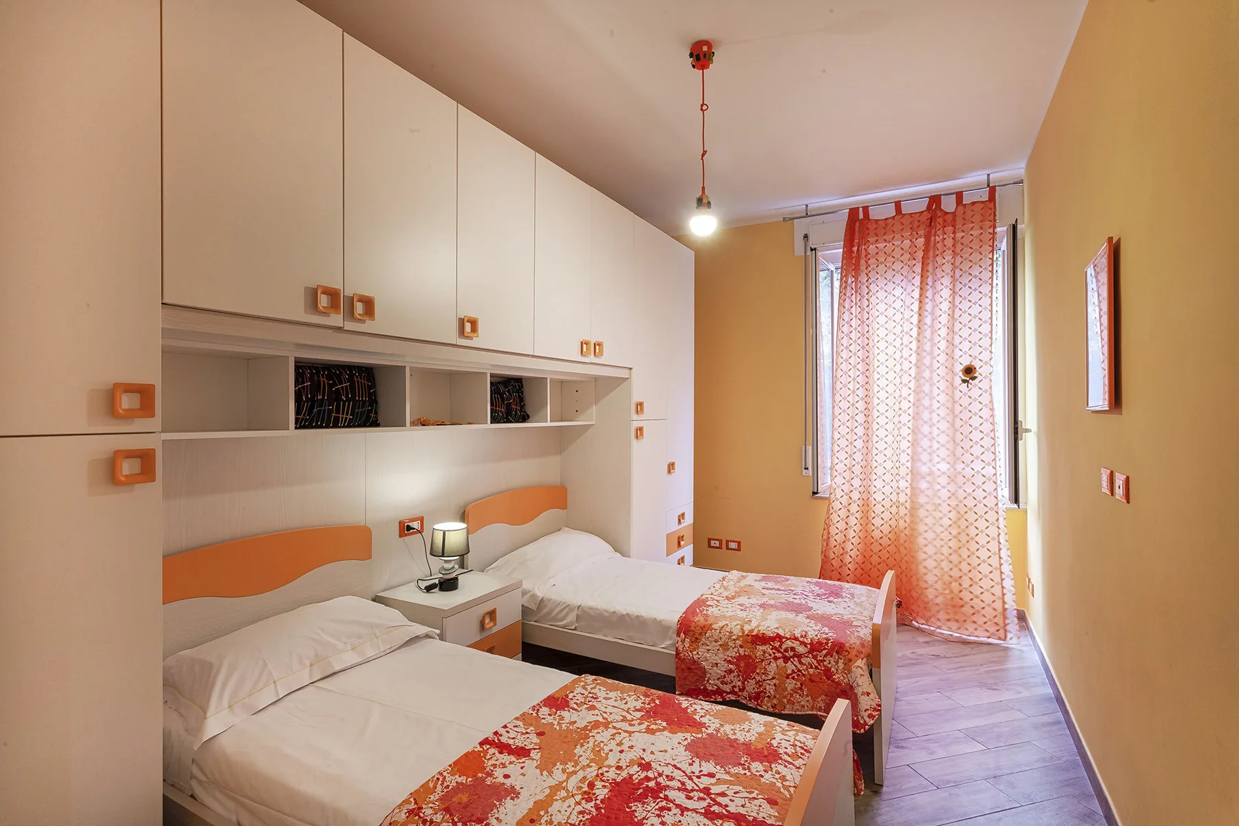 A bedroom with two beds and orange patterned curtains by the window.