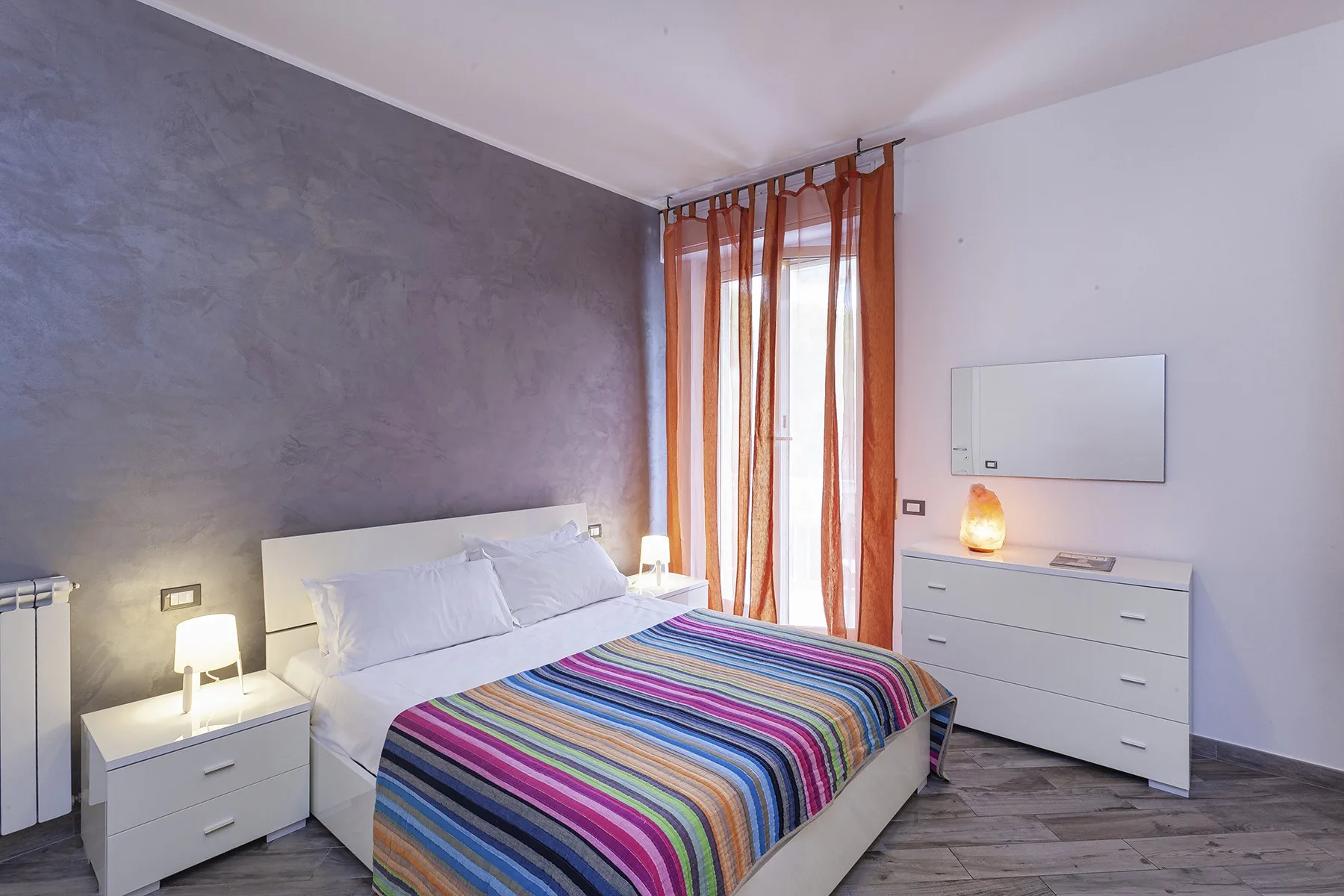 A bedroom with a bed covered by a colorful striped blanket and tall curtains.