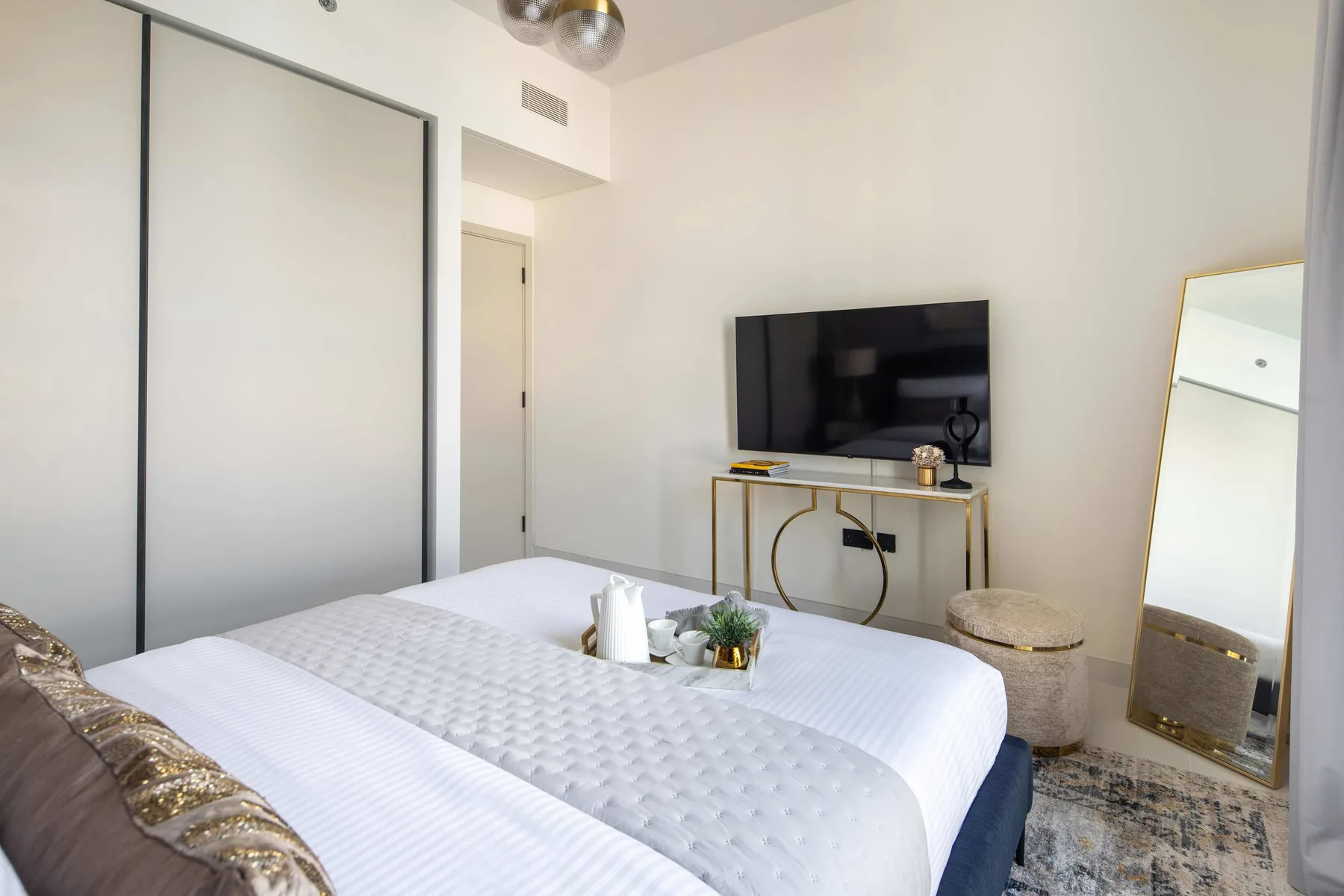 Un dormitorio luminoso con una cama bien tendida, un televisor en la pared y un espejo de cuerpo entero.