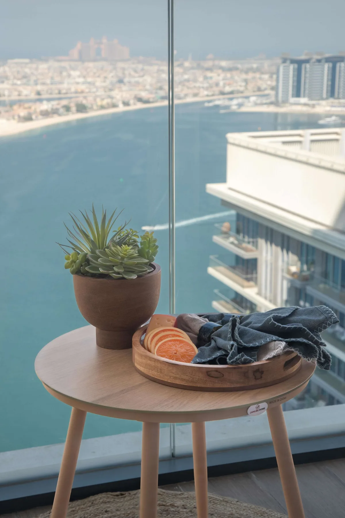Una mesa redonda pequeña con una planta en maceta y una bandeja está junto a una ventana con vistas al agua.
