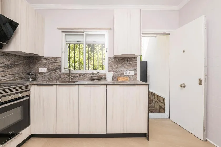 A bright kitchen with pale cabinets, dark countertops, and a window above the sink.