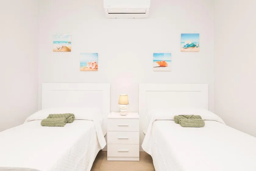 A bright bedroom with two beds, beach-themed pictures, and a wall-mounted air conditioner.