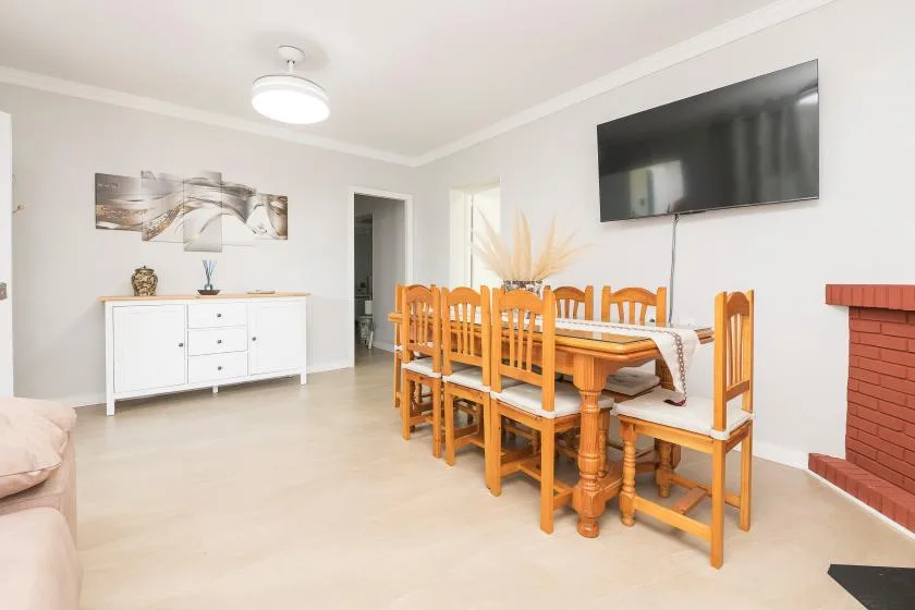 A bright dining room with a wooden table and chairs beneath a wall-mounted television.