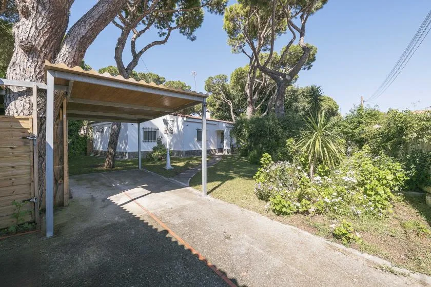 A covered driveway sits beside a white building, surrounded by trees and shrubs.