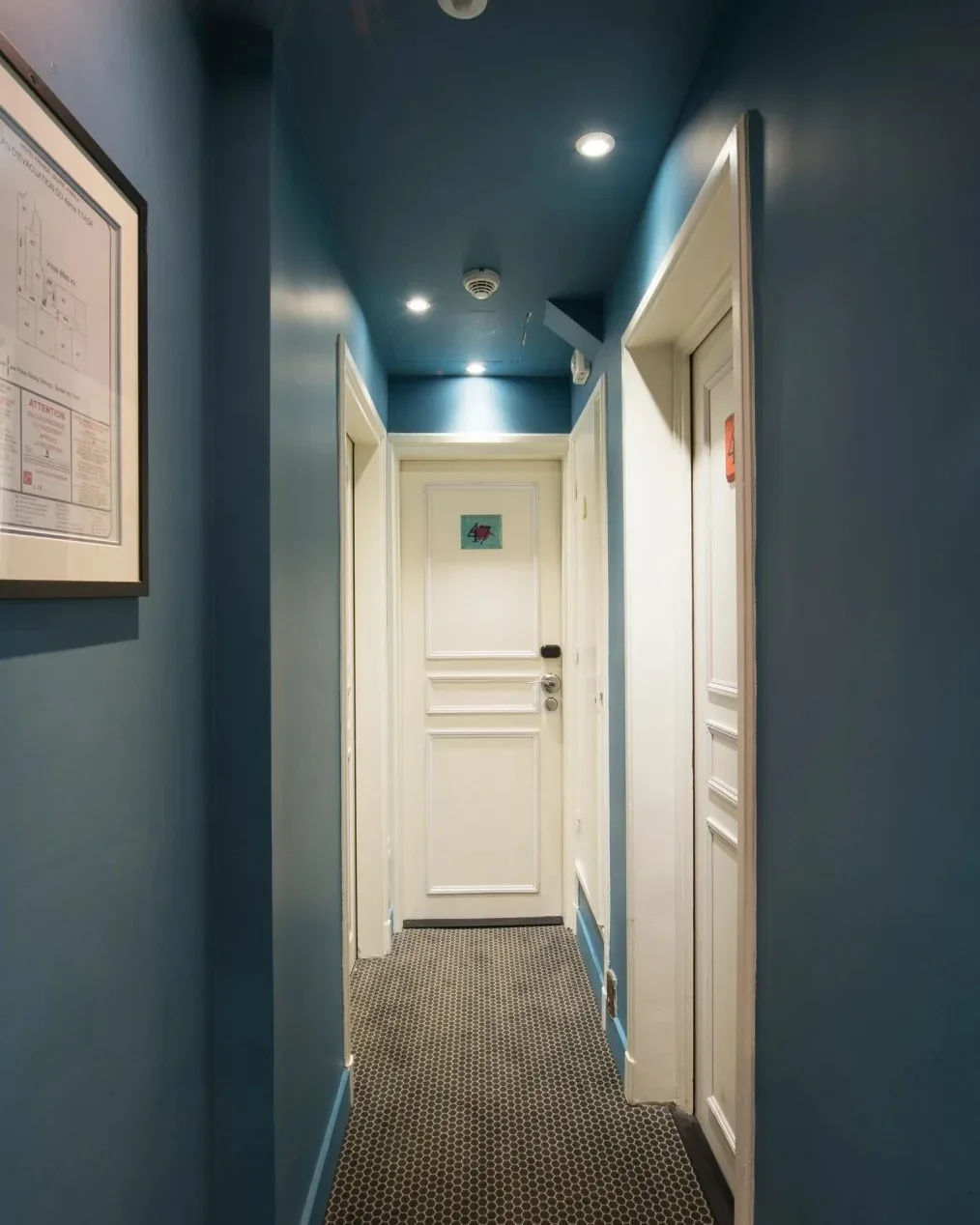 Pasillo de hotel decorado en azul, con puertas blancas y un cuadro en la pared.