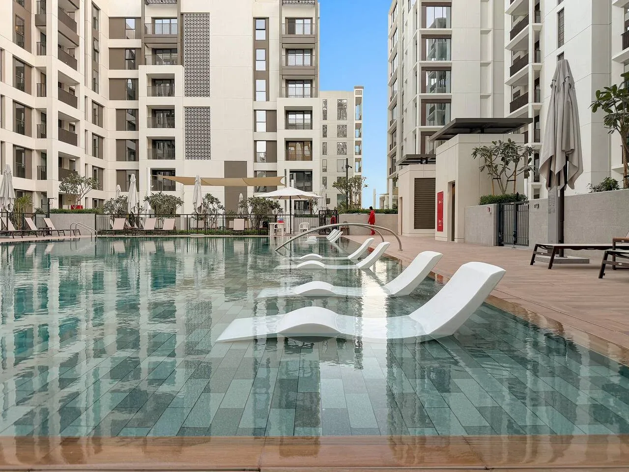 An outdoor swimming pool sits between modern apartment buildings, with submerged loungers.