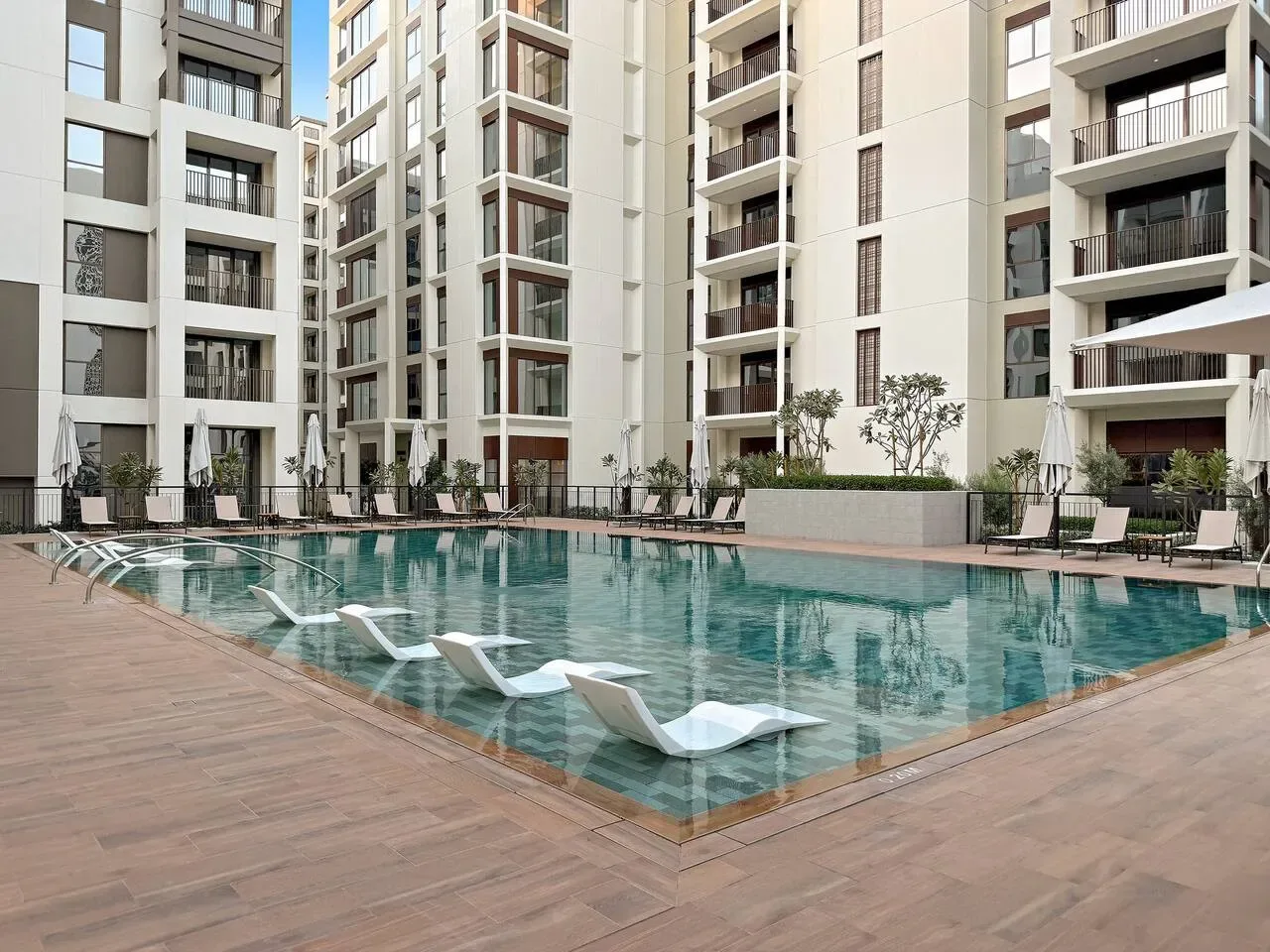 An outdoor swimming pool sits among tall apartment buildings, with lounge seating along the deck.