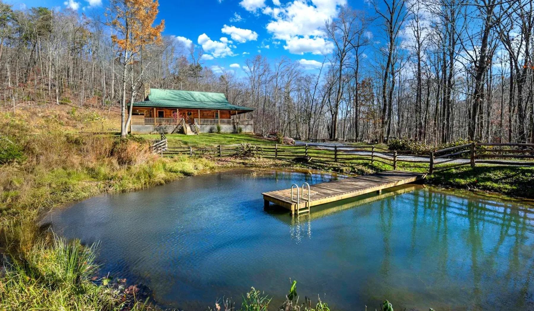 A pond with a small dock sits beside a wooded hillside and cabin.