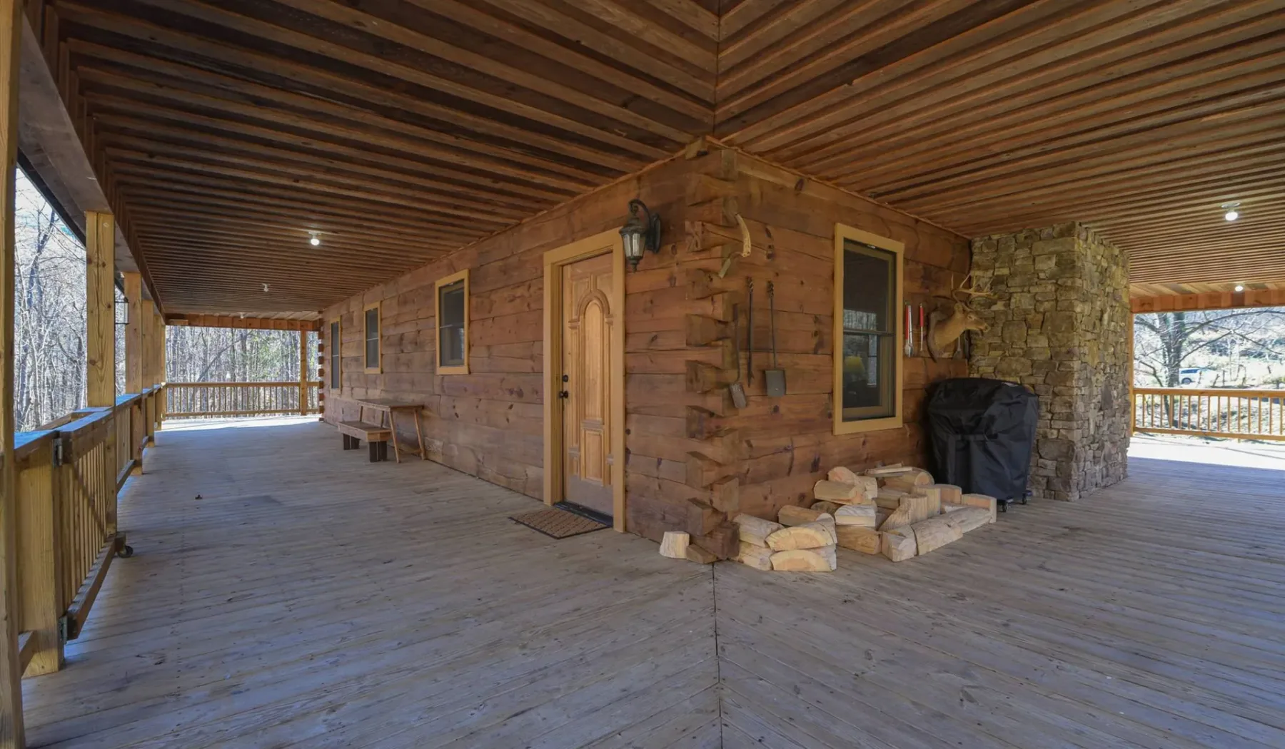 A covered wooden porch surrounds a rustic cabin, with a stone chimney beside the entrance.