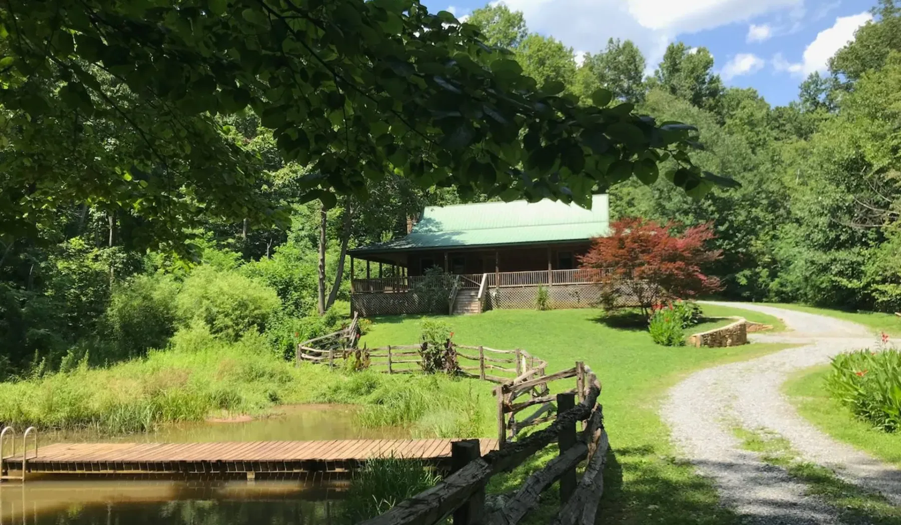 A rustic cabin sits beside a gravel drive, surrounded by green trees and lawn.