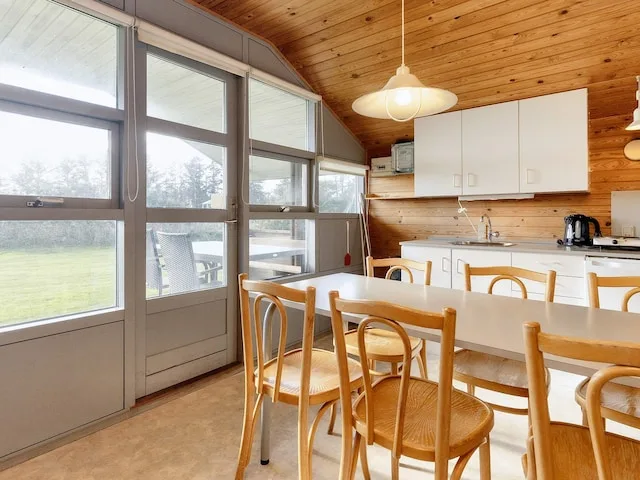 A dining area with wooden chairs beside large windows and a kitchen.