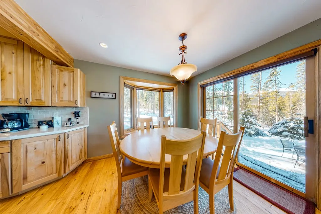A dining area with a wooden table and chairs beside large windows overlooking a snowy landscape.