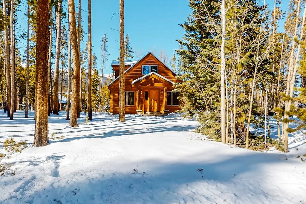 A wooden cabin stands among snow-covered trees beneath a clear blue sky.