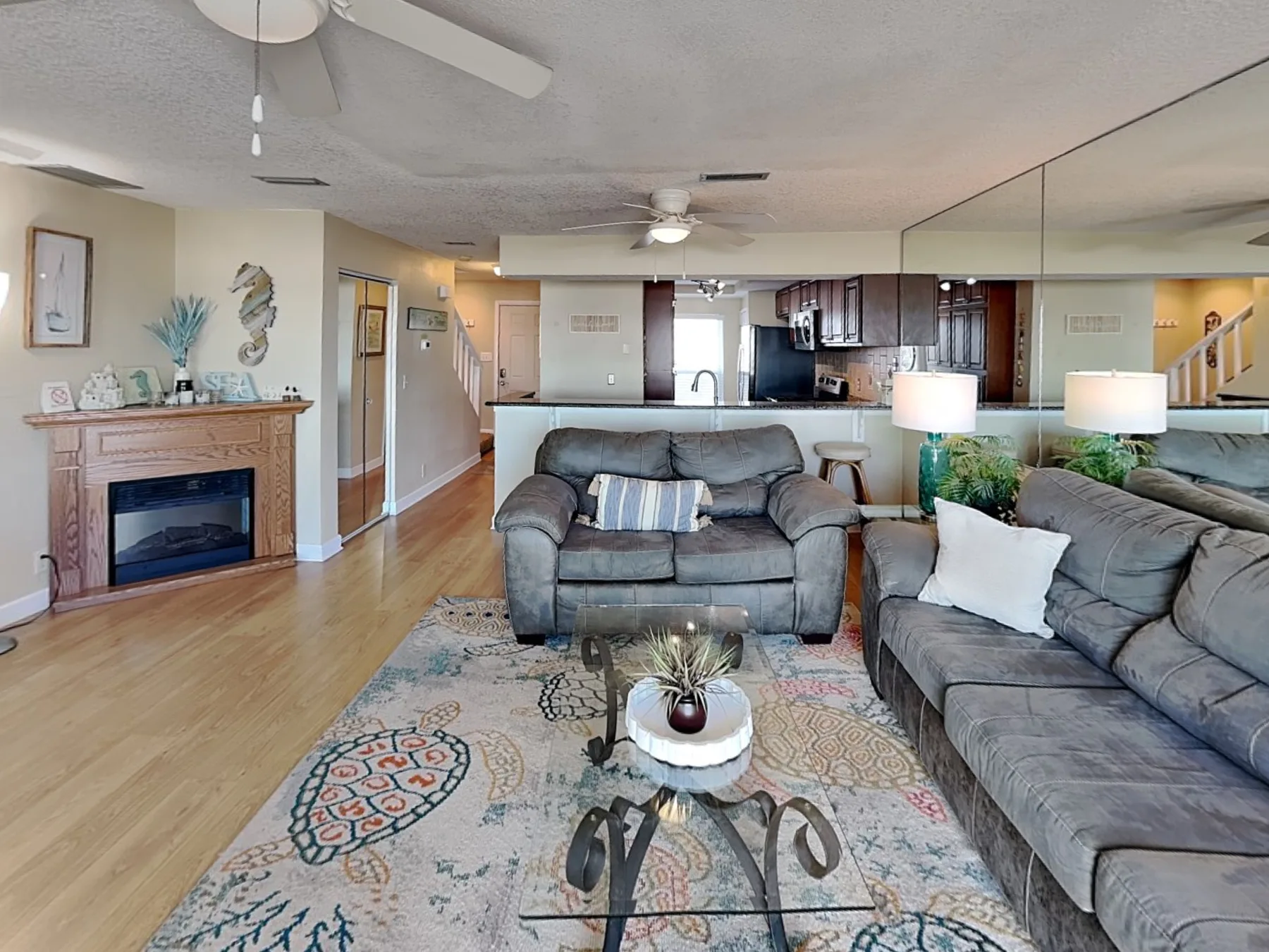 A living room with gray seating, a patterned rug, and an open kitchen behind it.