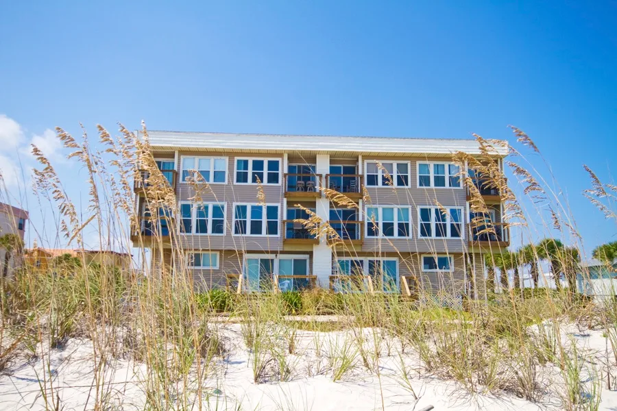 A multi-story beachfront building stands behind dune grass on a sunny day.