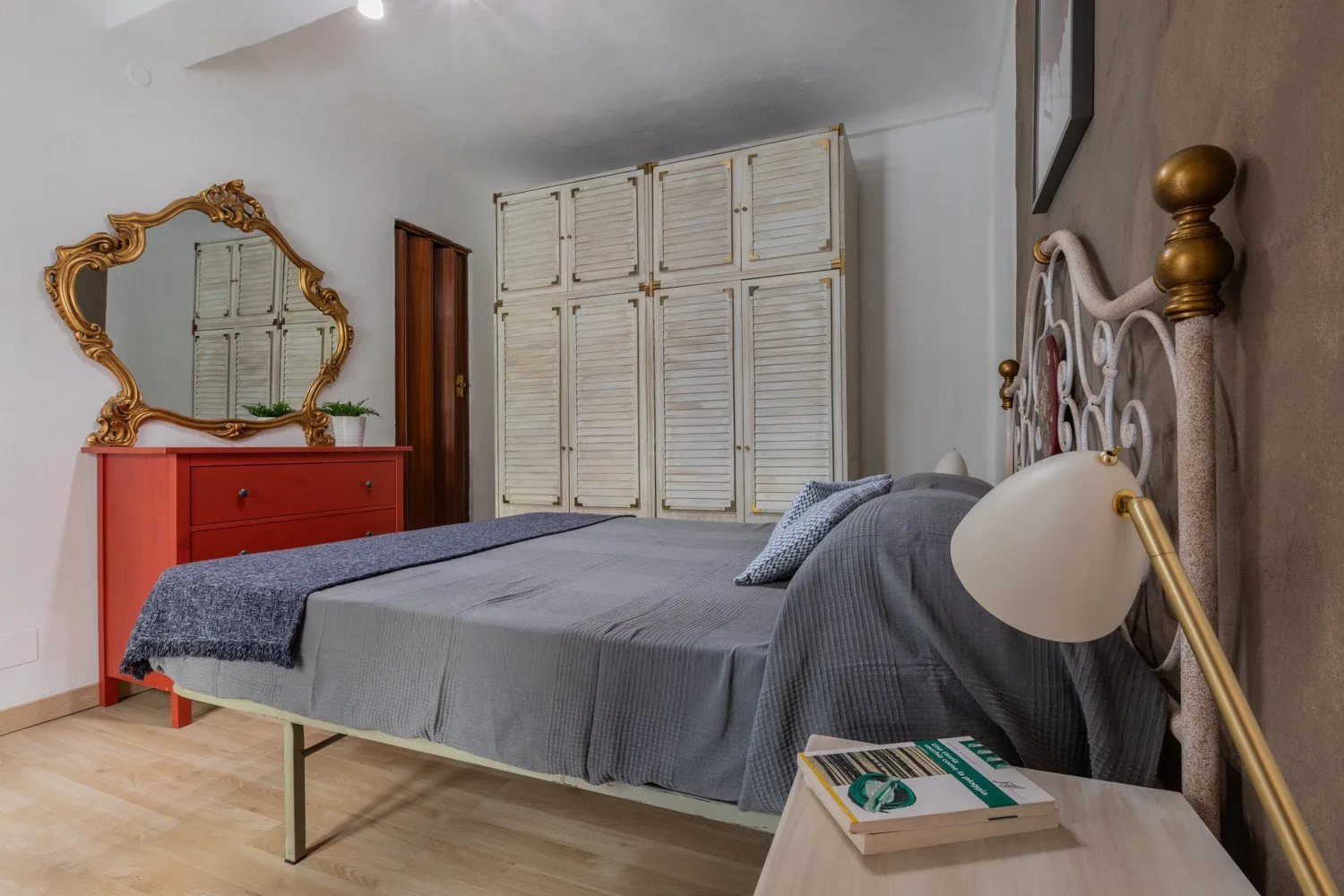 Un dormitorio con una cama cubierta de gris, persianas blancas y una cómoda roja.