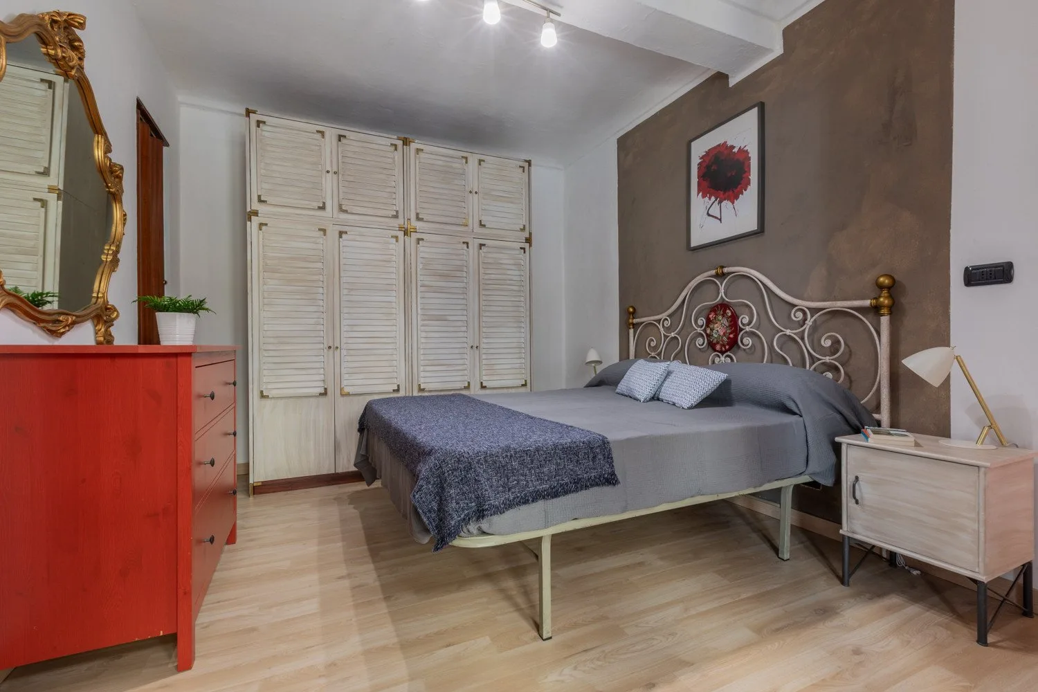 Un dormitorio con una cama de metal, ventanas con persianas y suelo de madera.