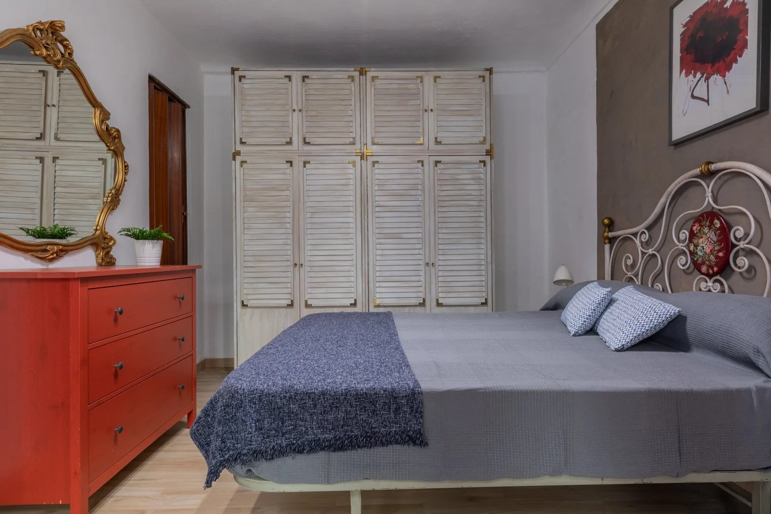 Un dormitorio con una cama gris, puertas blancas de armario con persianas y una cómoda roja.