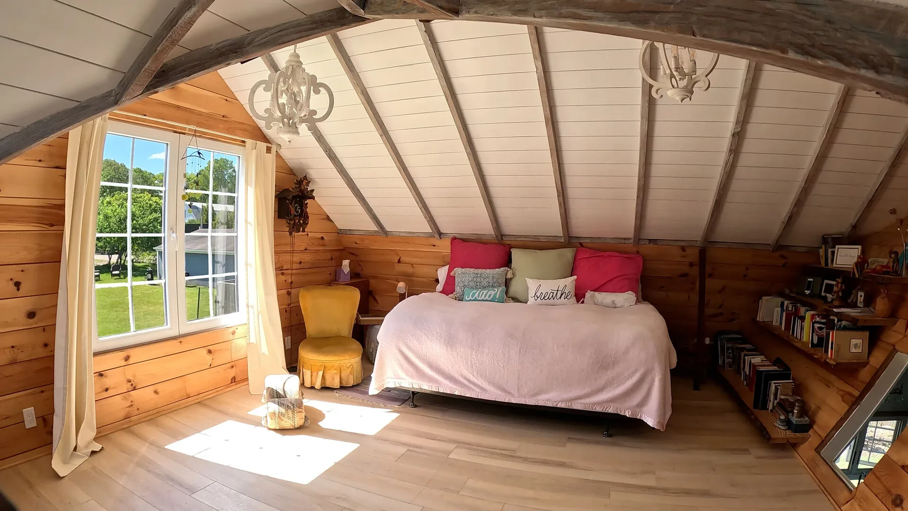 Dormitorio soleado con una cama bajo un techo inclinado y una ventana grande.