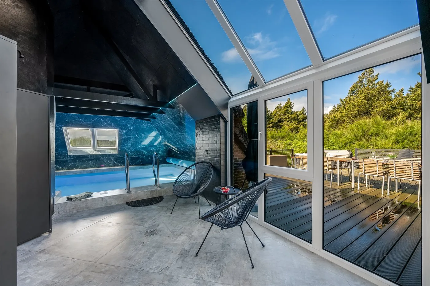 Sala con piscina cubierta y grandes ventanales con vistas a una terraza de madera.
